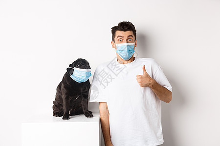 Covid19 动物和检疫概念 在医疗面具中装有搞笑的年轻人和小狗的照片 拥有者在认可或类似时露出拇指 白色背景社交宠物工作室促图片