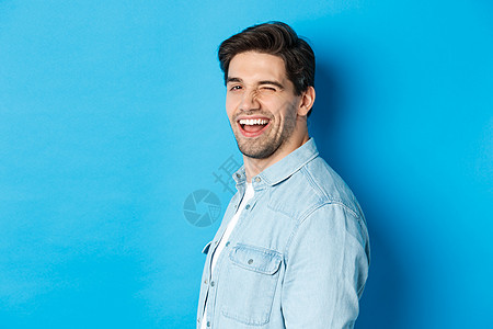 快乐和自信的人将头转向镜头 眨着眼睛笑着微笑 站在蓝背景上蓝色黑发情绪技术男性闲暇促销商业潮人广告图片