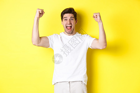 激动人心的男子获胜 举起手来庆祝 为团队赢得胜利和扎根 站在黄色背景之上的画面商业工作室成就快乐优胜者广告成人男性情绪手势背景图片