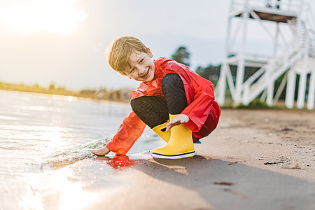 穿着红色雨衣和黄色橡胶靴的男孩在沙滩上玩水 穿着防水外套的小学生在海上接触水 孩子在岸边玩海浪海岸靴子支撑海洋波浪天气旅行海滩乐图片