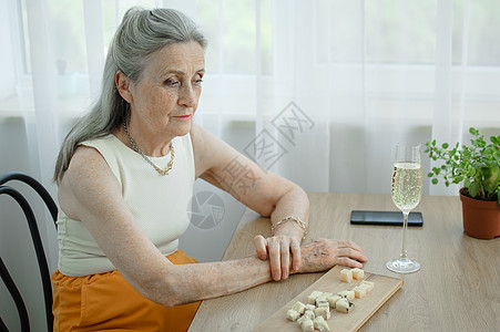 美丽的老祖母 头发灰白 脸上满是皱纹 坐在家里的桌子旁 窗户背景是一杯香槟 母亲节 退休快乐木板闲暇酒精有钱人情感饮料舒适老年幸图片