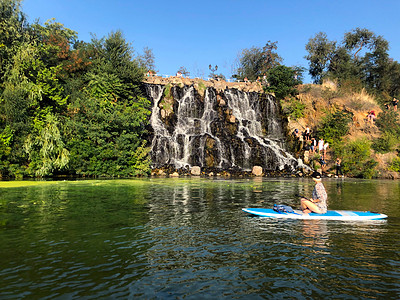 女孩在瀑布附近游泳 在河边休息 一群人靠近人工瀑布 瀑布周围绿树成荫 蓝天无云图片