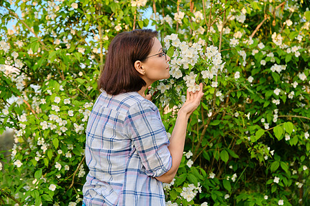 一个微笑的中年妇女 在一个开花的茉莉花丛旁的肖像茉莉花成人福利衬套衬衫花朵女性花园女士眼镜图片