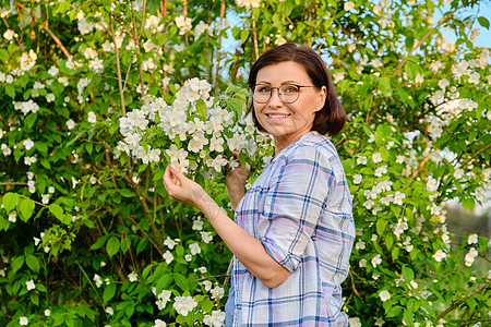 一个微笑的中年妇女 在一个开花的茉莉花丛旁的肖像女性日落衬套成人闲暇衬衫花园花朵福利眼镜图片
