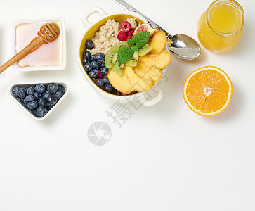 盘子里放着燕麦片和水果 半个成熟的橙子和鲜榨果汁放在透明玻璃瓶中 蜂蜜放在白桌上的碗里 健康早餐谷物小吃甜点勺子燕麦粮食果汁桌子图片