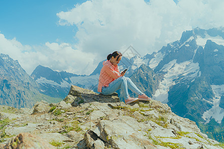 在山中使用智能手机的女性旅行者 在阳光明媚的日子里 全身女人坐在岩石上 在多云的天空中浏览智能手机旅行手表天气冒险冲浪社交旅游系图片