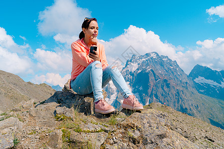 在山中使用智能手机的女性旅行者 在阳光明媚的日子里 全身女人坐在岩石上 在多云的天空中浏览智能手机社交远足工具定位农村旅游石头探图片