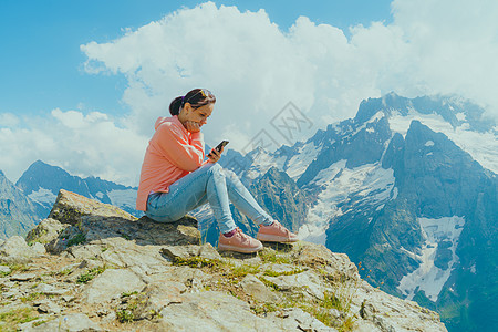 在山中使用智能手机的女性旅行者 在阳光明媚的日子里 全身女人坐在岩石上 在多云的天空中浏览智能手机社交天气手表导航互联网定位农村图片