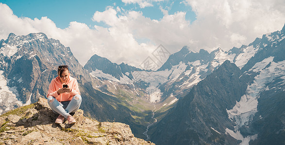 在山中使用智能手机的女性旅行者 在阳光明媚的日子里 全身女人坐在岩石上 在多云的天空中浏览智能手机社交冒险假期农村晴天媒体顶峰旅图片
