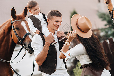 白衣家庭与儿子一起站在两匹美丽的自然马旁 一对有孩子的时装夫妇被用马拍下照片 a 一名男子帽子男人男生幸福孩子们农村白色女士农场图片
