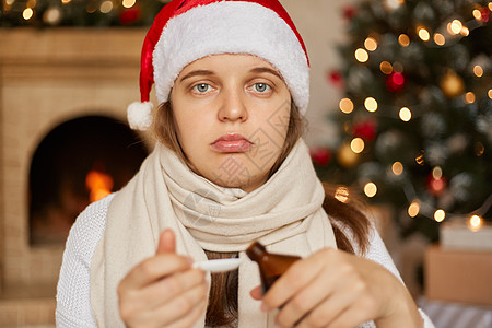 戴着圣诞帽 因咳嗽 因病毒或感染而生病的糖浆的年轻白人女性 用不安的表情看着镜头 裹着战巾 在装饰着新年装饰的房间里摆姿势图片