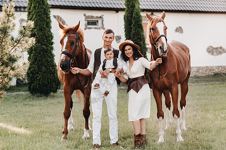 白衣家庭与儿子一起站在两匹美丽的自然马旁 一对有孩子的时装夫妇被用马拍下照片 a 一名男子母亲娱乐动物日落牛仔闲暇白色帽子农村牧图片