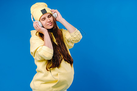 一个穿黄色夹克和耳机的怀孕女孩 站在蓝色背景上运动衫立体声笑声帽子休息歌曲女士衣服音乐乐趣图片