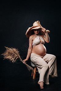 在黑色背景的工作室里 戴着草帽 穿着米色衣服 手里拿着花束的孕妇怀孕身体母亲女孩场地成人稻草大肚子腹部幸福图片