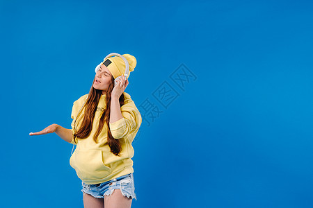 一个穿黄色夹克和耳机的怀孕女孩 站在蓝色背景上短裤歌曲牛仔裤腹部唱歌闲暇音乐乐趣女士立体声图片