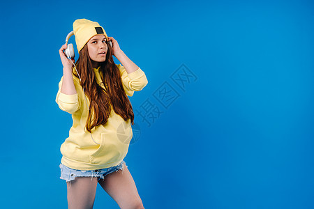 一个穿黄色夹克和耳机的怀孕女孩 站在蓝色背景上音乐乐趣唱歌立体声笑声运动衫情感微笑休息女士图片
