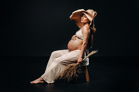 在黑色背景的工作室里 戴着草帽 穿着米色衣服 手里拿着花束的孕妇肚子帽子孩子大肚子成人花朵母亲场地母性裤子图片