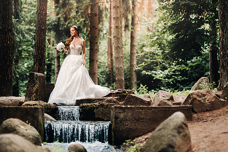 一位身着白色连衣裙 戴着手套 手捧花束的优雅新娘站在森林的溪流旁 享受着大自然 自然公园里穿着婚纱和手套的模特 白俄罗斯裙子男人图片