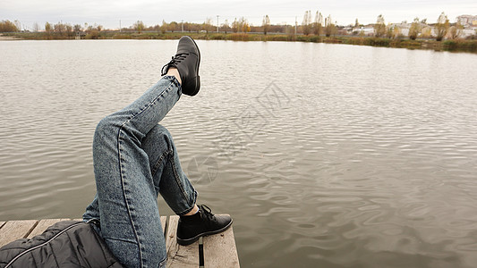 一个穿着蓝色牛仔裤的女孩坐在湖边 一位年轻女子正在河边休息 坐在一座木桥的边缘 双腿在木码头的湖边放松 人们在休息女士海岸乐趣假图片