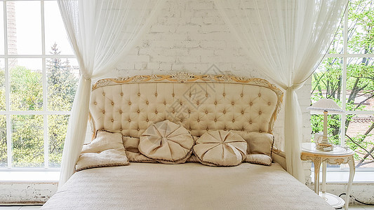 一张舒适的白色木床靠近一扇大窗户 房间里挂着漂亮的窗帘 金框现代咖啡桌 光线优雅 白色工作室内部配有一张大床用品风格床头奢华羽绒图片