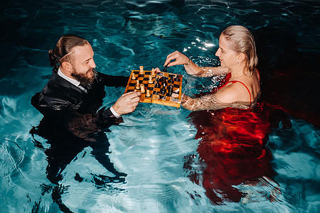 一个穿西装的男人和一个穿红裙子的女孩 在游泳池的水上下棋礼服闲暇成年人象棋运动温泉浴消遣娱乐玩家套装图片