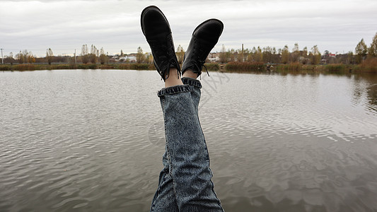 一个穿着蓝色牛仔裤的女孩坐在湖边 一位年轻女子正在河边休息 坐在一座木桥的边缘 双腿在木码头的湖边放松 人们在休息情绪女士鞋类运图片