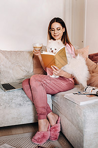 一个穿着睡衣的女孩在家里和她的狗 Spitzer 一起看书 狗和它的主人正在沙发上休息并看书 家务享受眼镜工具动物女性小狗杂志放图片