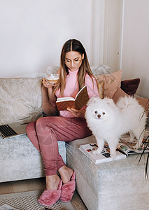 一个穿着睡衣的女孩在家里和她的狗 Spitzer 一起看书 狗和它的主人正在沙发上休息并看书 家务动物成人房间微笑乐趣公寓宠物工图片