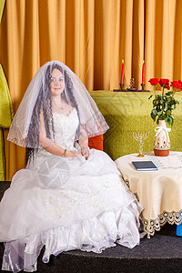 一位穿着白色礼服的犹太新娘 她的脸被面纱蒙上面纱 坐在餐桌旁 在教堂仪式前送花图片
