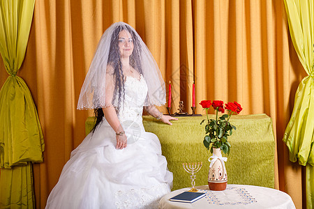 一位幸福的犹太新娘穿着蓬松的白色连衣裙 脸上蒙着面纱 在 chupa 仪式前的房间里站在桌子旁图片