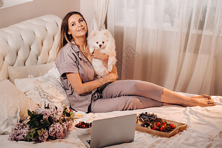 一个穿着睡衣的女孩晚上坐在床上 她的白狗看着笔记本电脑吃着糖果 家里有一只斯皮策犬的女孩躺在床上食物休息枕头苏醒工作早餐电影互联图片