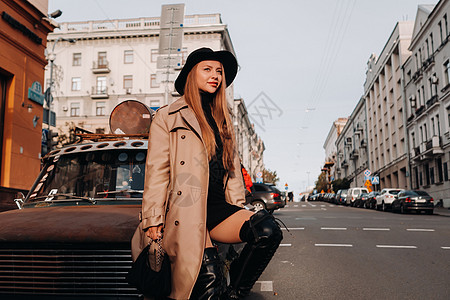 日落时分 城市街道上一位身着米色外套 头戴黑色帽子的时尚年轻女子坐在汽车的引擎盖上 女性街头时尚 秋装 都市风太阳镜手提包街道城图片