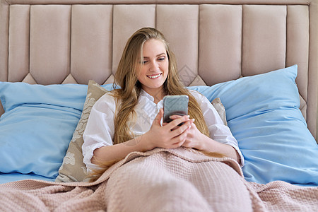 年轻美丽 笑着的金发美女 家里床上躺着智能手机图片