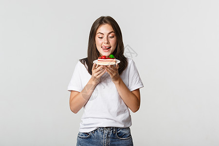 被诱惑的美丽女孩舔嘴唇看蛋糕的肖像女性学生女士女朋友成人享受黑发手势青年广告图片