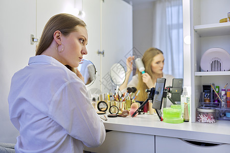 年轻女士坐在化妆桌前 照着镜子做化妆和发型化妆台互联网皮肤技术调色板女孩博主金发女郎头发造型图片