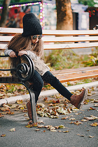 4-5 岁的时尚女婴戴着针织帽 太阳镜 靴子 毛皮大衣坐在公园的长椅上 看着相机 秋天秋天的季节图片