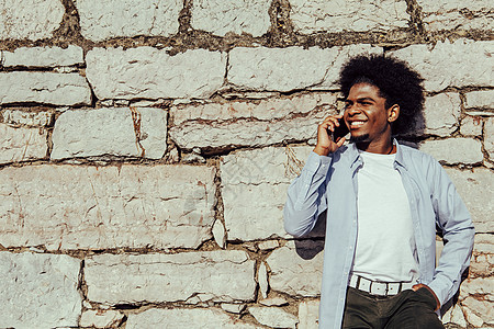 非洲年轻人在手机上交谈时 仰靠墙壁的快乐成人微笑享受男性细胞电话青年倾斜技术图片