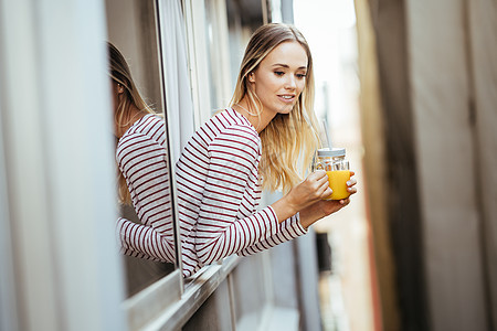 年轻女子喝着一杯天然橙汁 从家窗外倾斜而下 从家里走出来食物玻璃稻草成人饮食女性窗户头发橙子水果图片