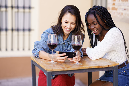 两个女人一边喝着酒一边看智能手机友谊玻璃女孩头发黑发辫子女性酒吧发型微笑图片