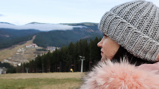 一位身穿粉红色毛皮夹克 头戴灰色针织帽的女山地游客的后视图 背景是群山 望着森林的秋季景观和多云的天空冒险旅行女士山脉娱乐旅游女图片