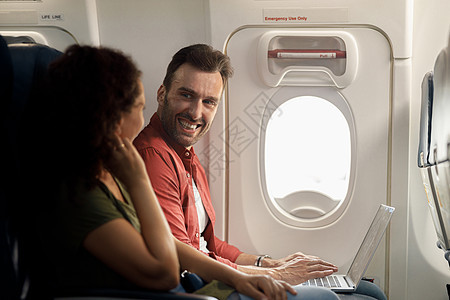 愉快的caucasian男子在用笔记本电脑坐在飞机上时微笑着他的妻子图片