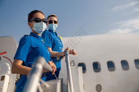 两名穿蓝制服的年轻空姐 太阳镜和防护面罩 在白天站在空中看着照相机 站立在空中背景图片