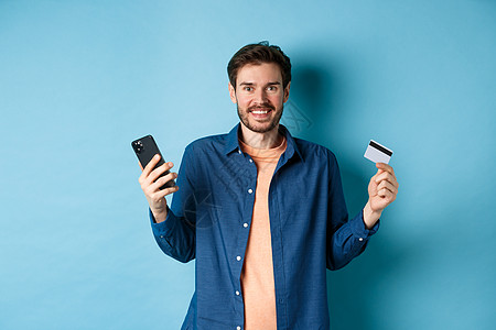 拥有智能手机和塑料信用卡 在线购物 站在蓝底的兴奋青年男子 在网上买东西以及以蓝底背景站立成功电话男人促销工作室发型胡须胡子技术背景图片