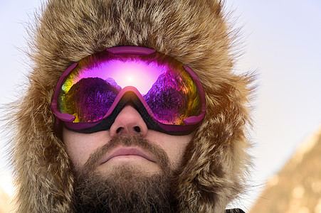 冬季雪山背景下 一位戴着滑雪面罩 戴着护目镜和一顶毛皮大老式帽子的留着胡须的快乐滑雪者的特写图片