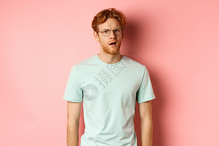 戴眼镜的帅气红发男子的画像看起来很困惑 张着嘴盯着镜头 什么都不懂 站在粉红色背景上冒充购物成人粉色头发男性广告眼镜红色工作室图片