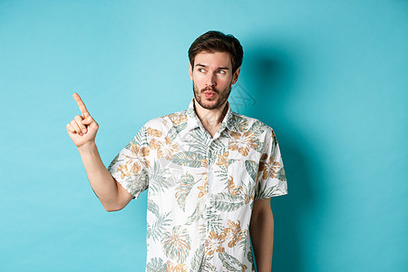 帅哥在度假时穿着夏季衬衫 指指点点地看着空荡荡的空间 站在蓝色背景上 脸上露出惊奇的表情工作室商业假期快乐旅游胡子旅行胡须男性广图片
