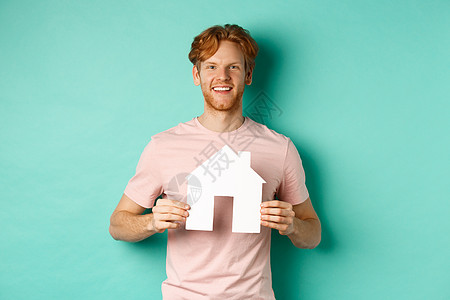 红色头发 穿T恤衫 露出纸屋剪裁和微笑的快乐 站在薄荷背景之上的年轻男子们胡子公寓学生机构发型房地产房子广告男性商业图片