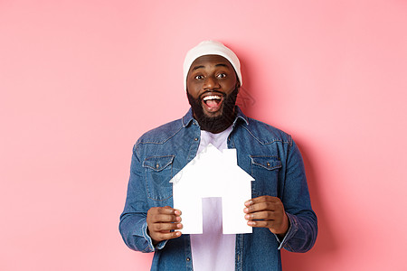 幸福快乐 笑笑的非洲美国人拥有房屋模型 兴奋地看着相机 寻找家园 站在粉红背景上站着 笑声胡须成人公寓胡子广告房地产帽子横幅房子图片