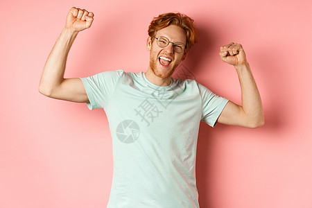 快乐的红头发年轻人看起来很开心 举起手来 举起拳头 庆祝成功 感觉像冠军 赢得胜利 站在粉红色的背景下男性胡须工作室胡子红色购物图片