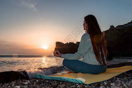 穿着蓝色牛仔裤和运动服的长头发的年轻女子在日落时分在海边的瑜伽垫上在户外练习 女性瑜伽健身程序 健康的生活方式 和谐与冥想海洋游图片